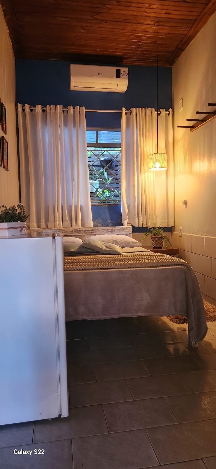 Cozy bedroom with bed, white curtains, potted plants, and wooden ceiling.